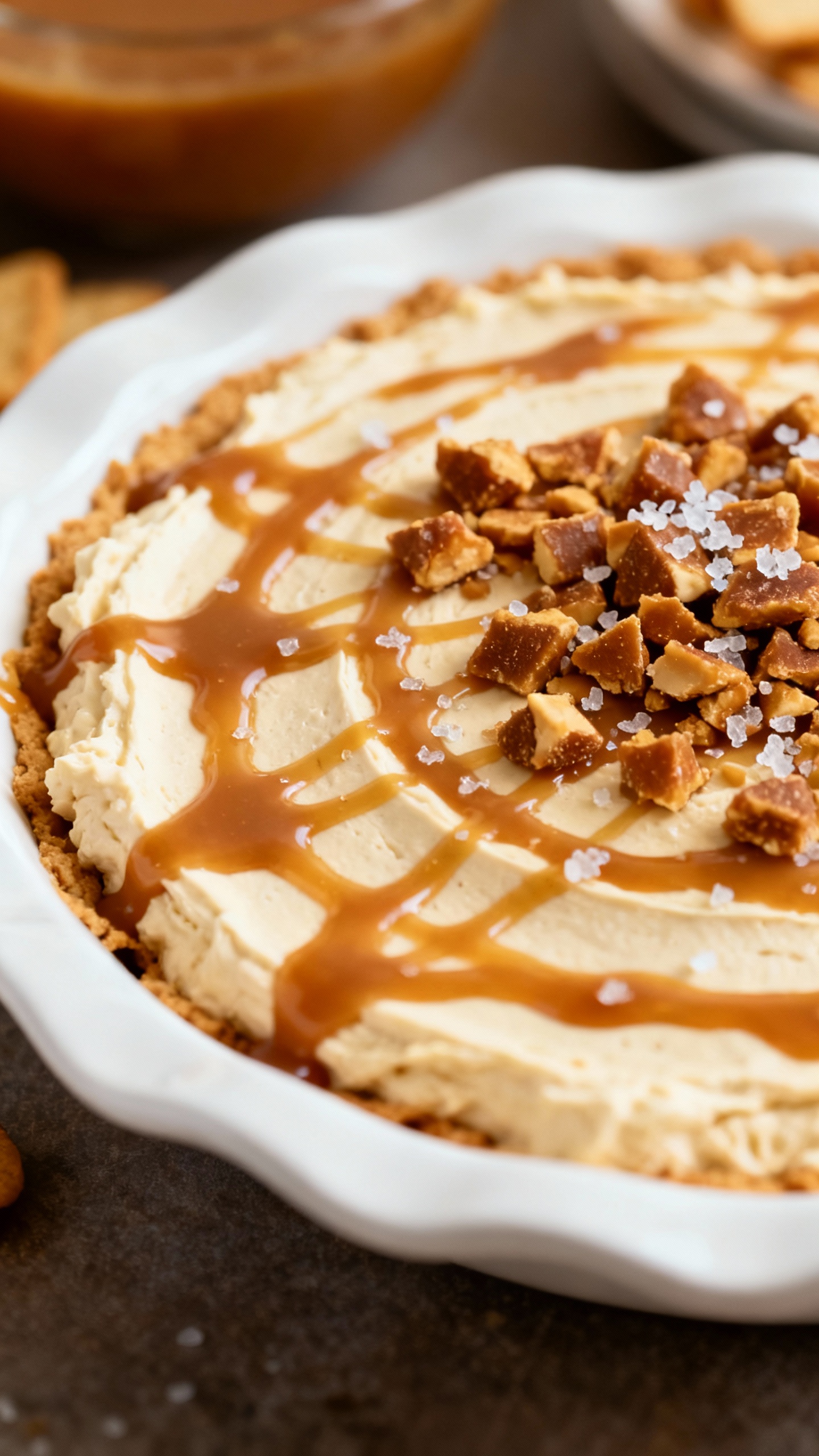 Closeup caramel-layered cream cheese dip in white pie plate, drizzled, topped with toffee bits and f