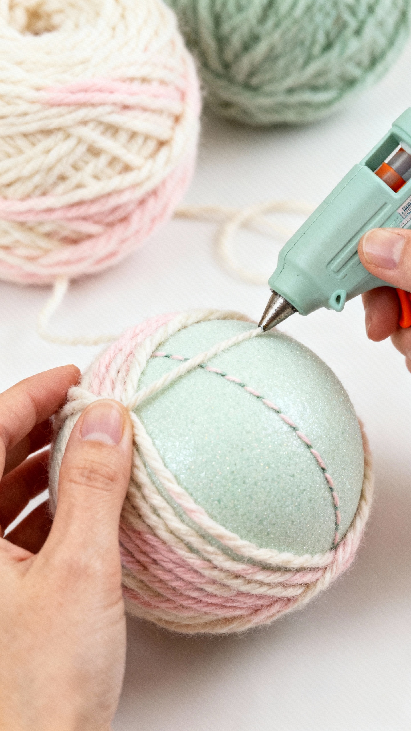 Hands wrapping chunky yarn around foam bauble, hot glue stringing, cream blush sage colors