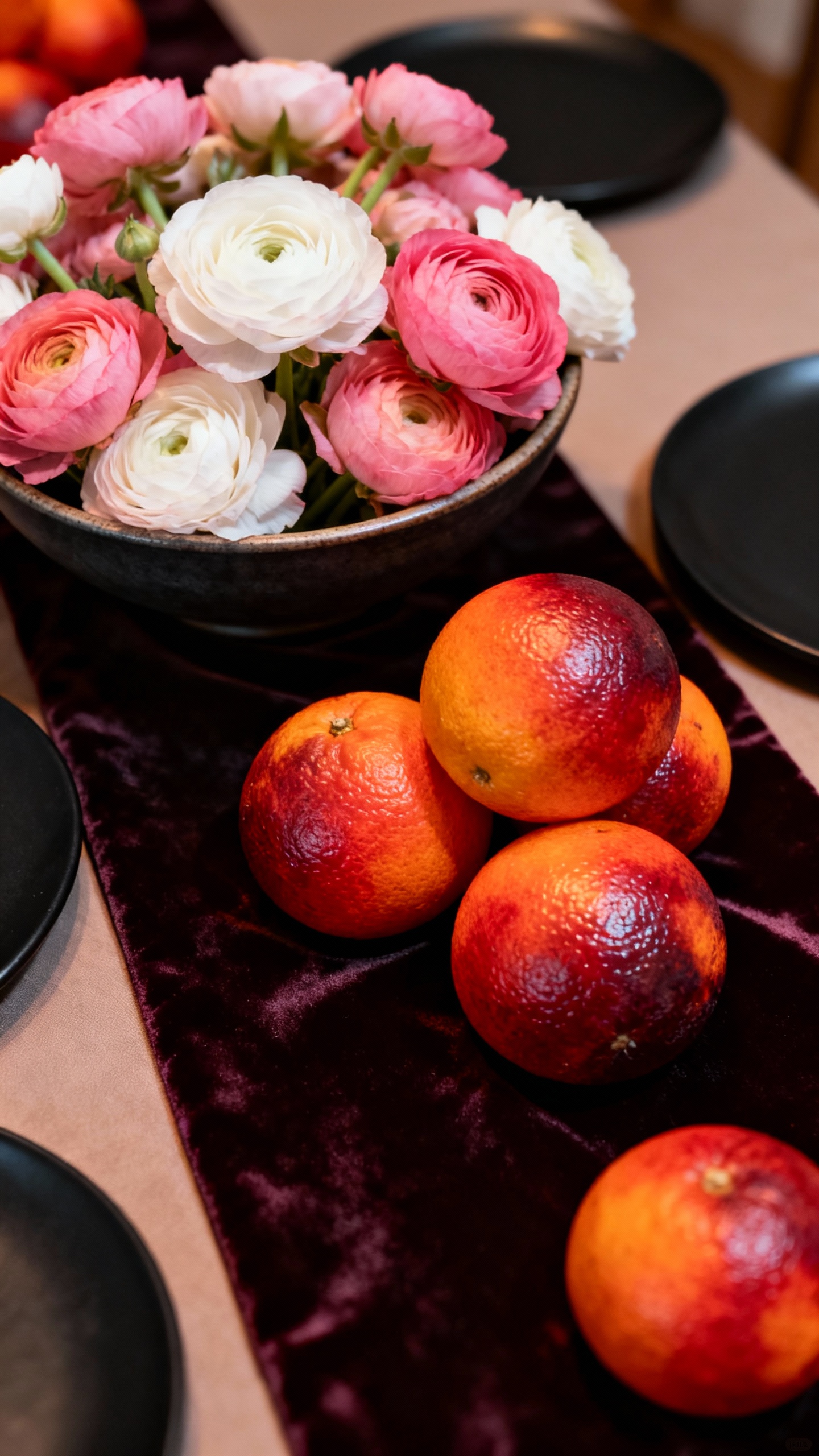 Low floral bowl with ranunculus beside blood oranges, velvet runner, matte black plates