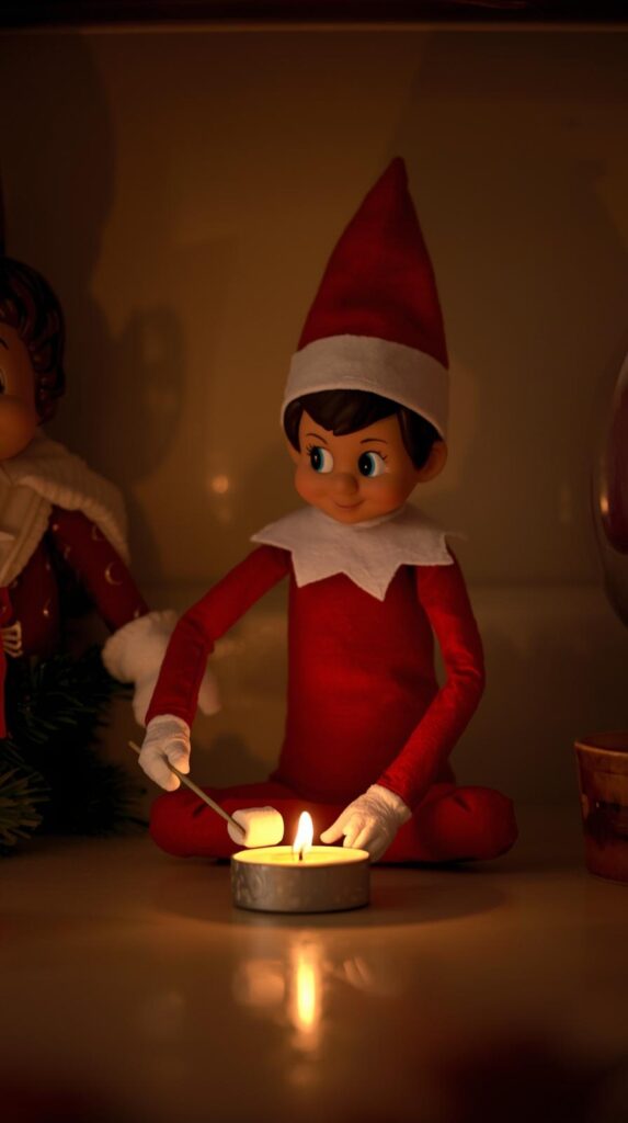 Photograph an Elf on the Shelf roasting mini marshmallows over a tealight candle on a kitchen counter. Warm lighting, cozy shadows, and playful holiday mood.